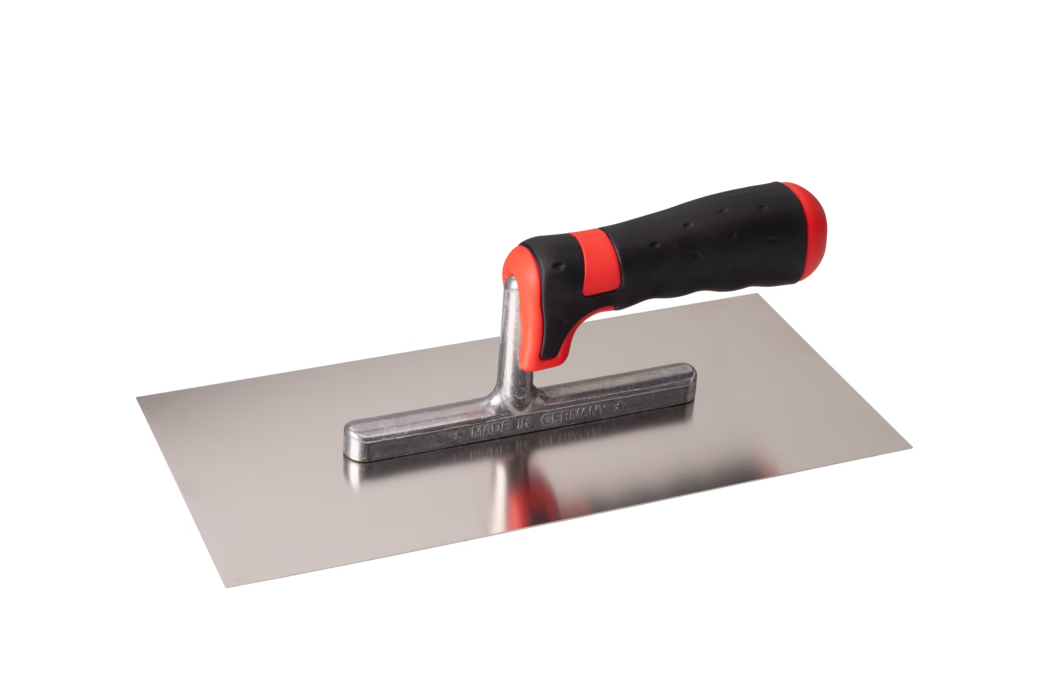 Truelle rectangulaire en acier inoxydable avec une lame lisse et plate et un court fût métallique supportant une poignée ergonomique noire et rouge à prise douce. La lame polie est réfléchissante et laisse apparaître une faible réflexion de la poignée ; le fût métallique est estampillé MADE IN GERMANY. La poignée présente des accents rouges à l'avant et à l'arrière et une surface en caoutchouc texturée pour une meilleure prise. L'outil est photographié isolé sur fond blanc, légèrement en contre-plongée. Utilisé pour appliquer et lisser le mortier, l'enduit ou le plâtre.